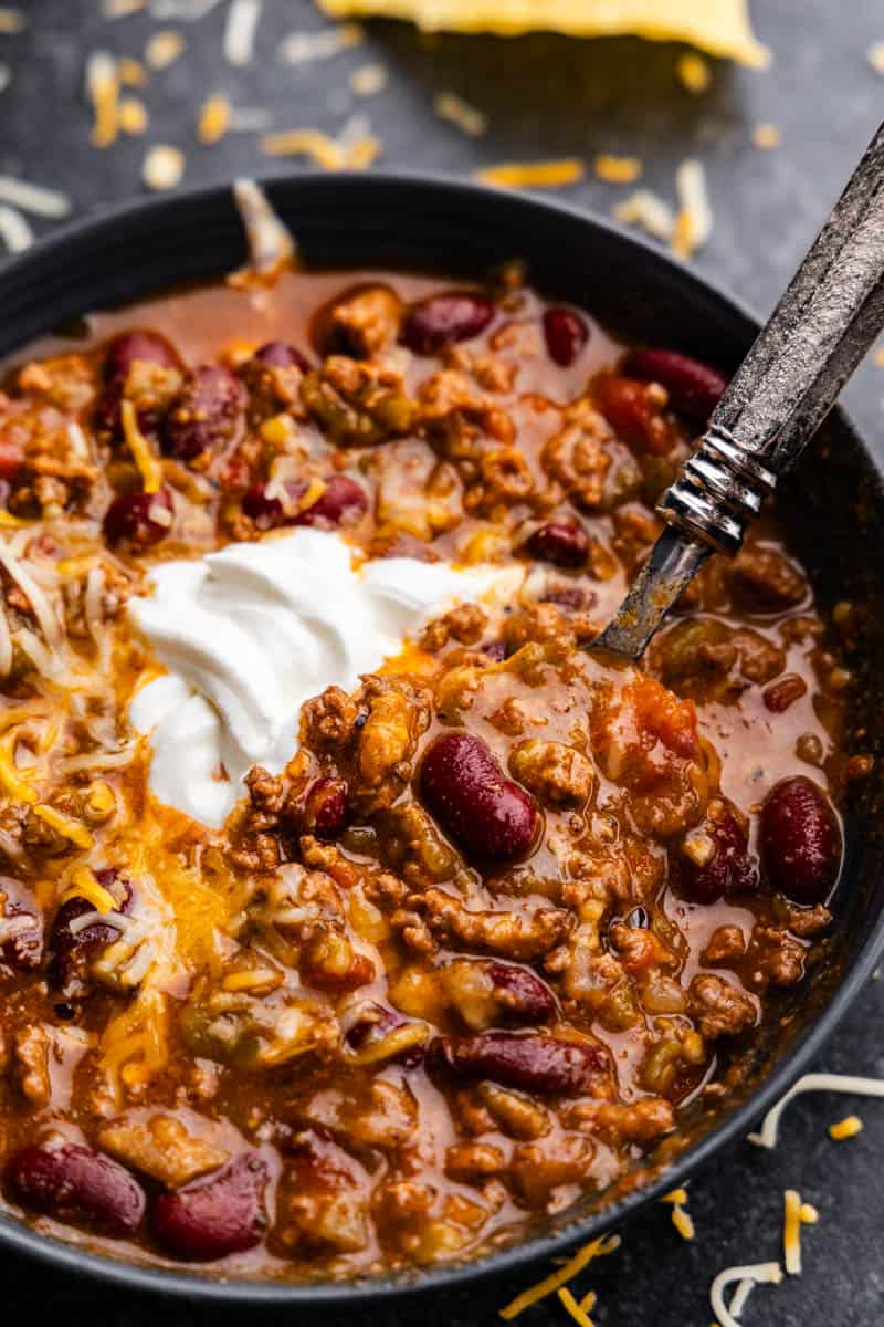 An overhead view of a bowl of no fuss chili topped with sour cream and cheddar cheese with a spoon set into the bowl for a bite.