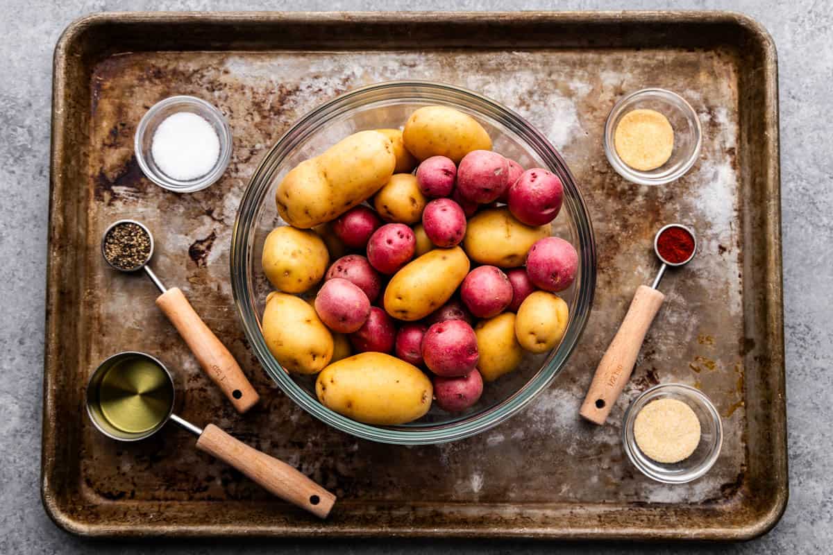 An overhead view of a sheet pan holding a bowl of mixed whole potatoes, and measuring cups and spoons with ingredients to make Breakfast Potatoes.