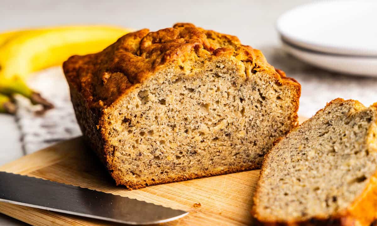 A loaf of banana bread set on a cutting board next to a knife, with a few slices laying in front of the loaf.