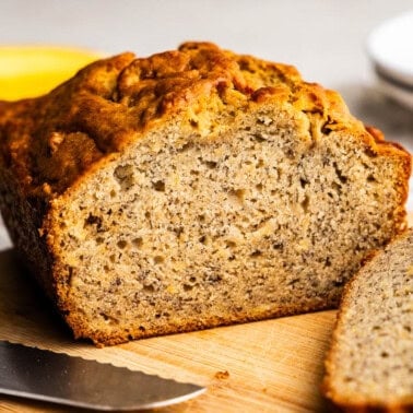 A loaf of banana bread set on a cutting board next to a knife, with a few slices laying in front of the loaf.