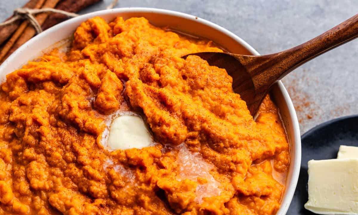 A close up overhead view of a large bowl of cinnamon honey butter mashed sweet potatoes. There's a pat of butter on top that's melting and a wooden spoon set in the bowl ready to serve.