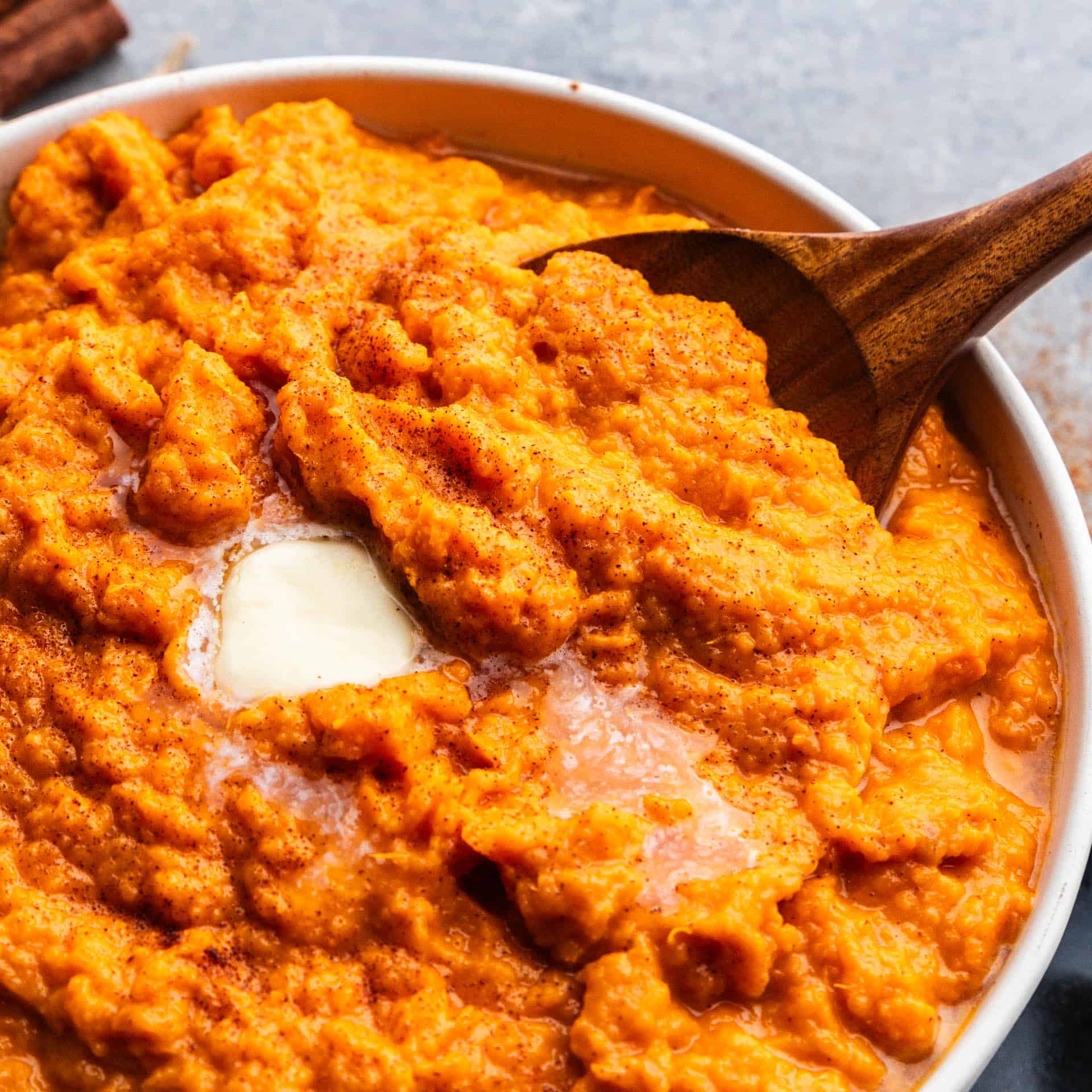 An overhead view into a bowl of cinnamon honey butter mashed sweet potatoes topped with a square of butter, with a wooden spoon set into the bowl for serving.