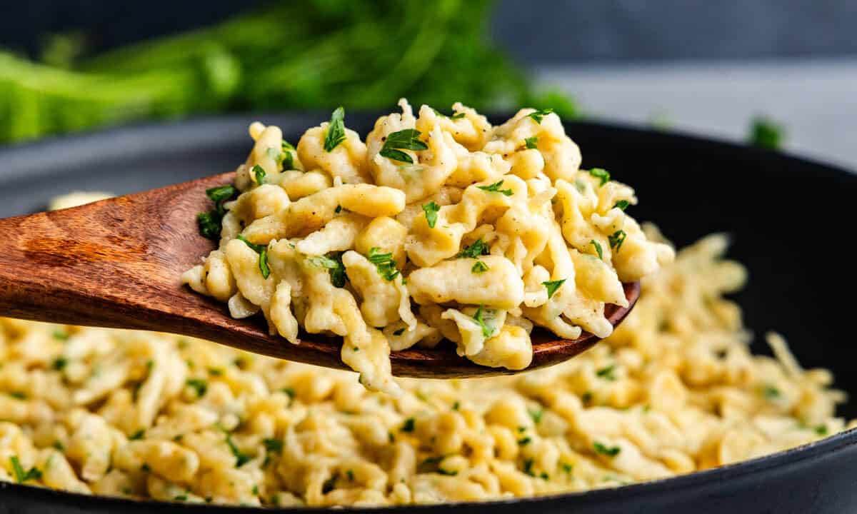 A side view of a wooden spoon holding a serving of German spaetzle above the skillet of the rest of the dish.