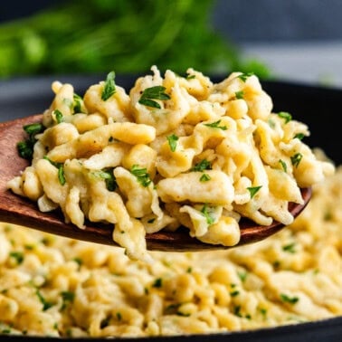 A side view of a wooden spoon holding a serving of German spaetzle above the skillet of the rest of the dish.