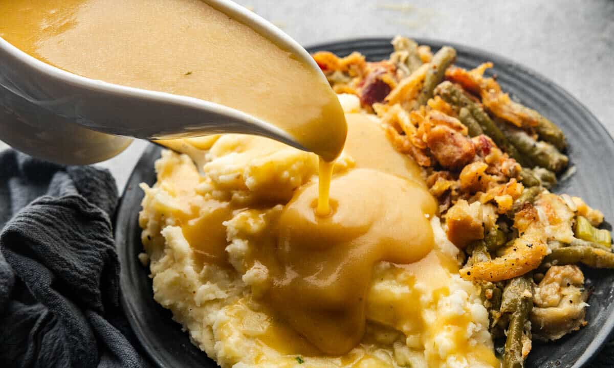 A gravy boat pouring gravy onto a plate of mashed potatoes and green bean casserole.