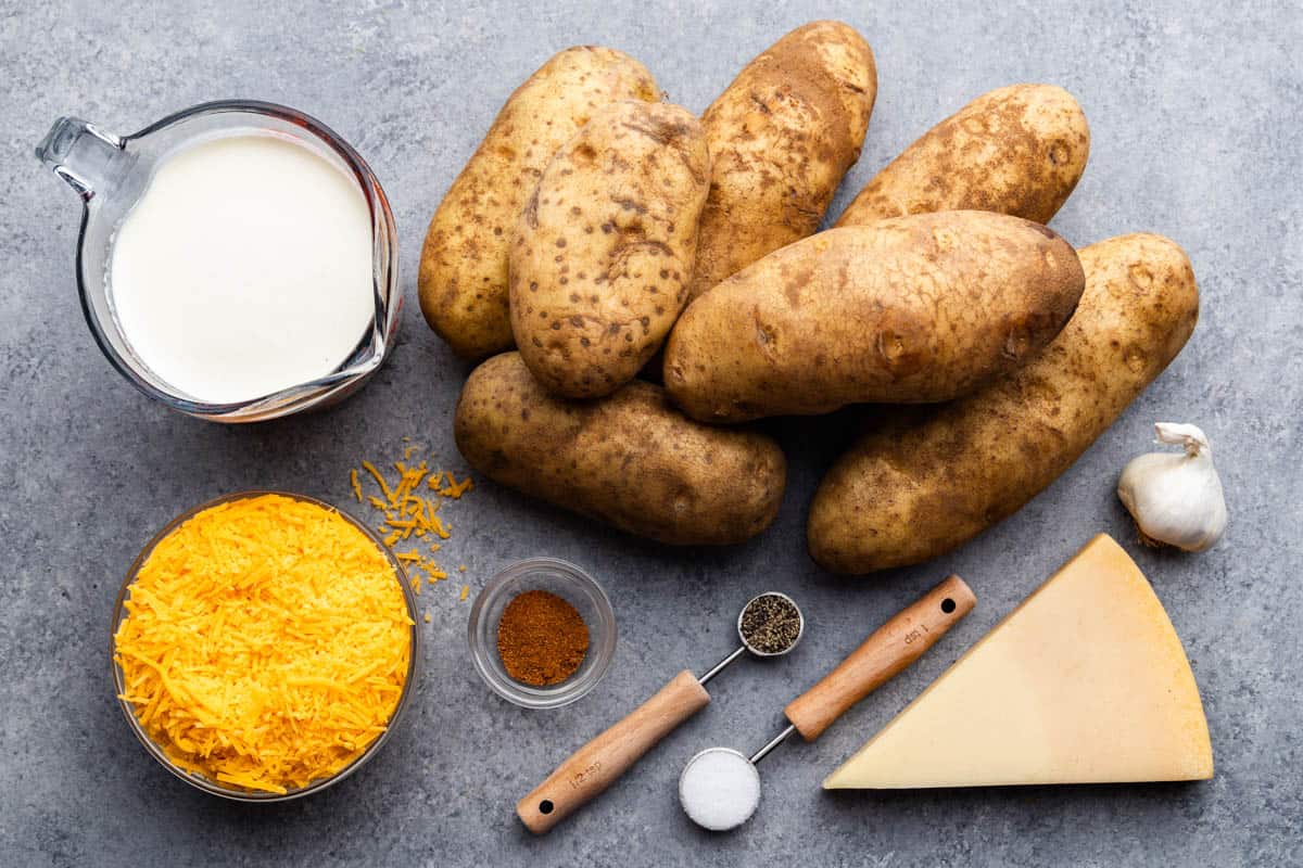 An overhead view of whole potatoes set next to measured wet and dry ingredients needed to make cheesy scalloped potatoes.