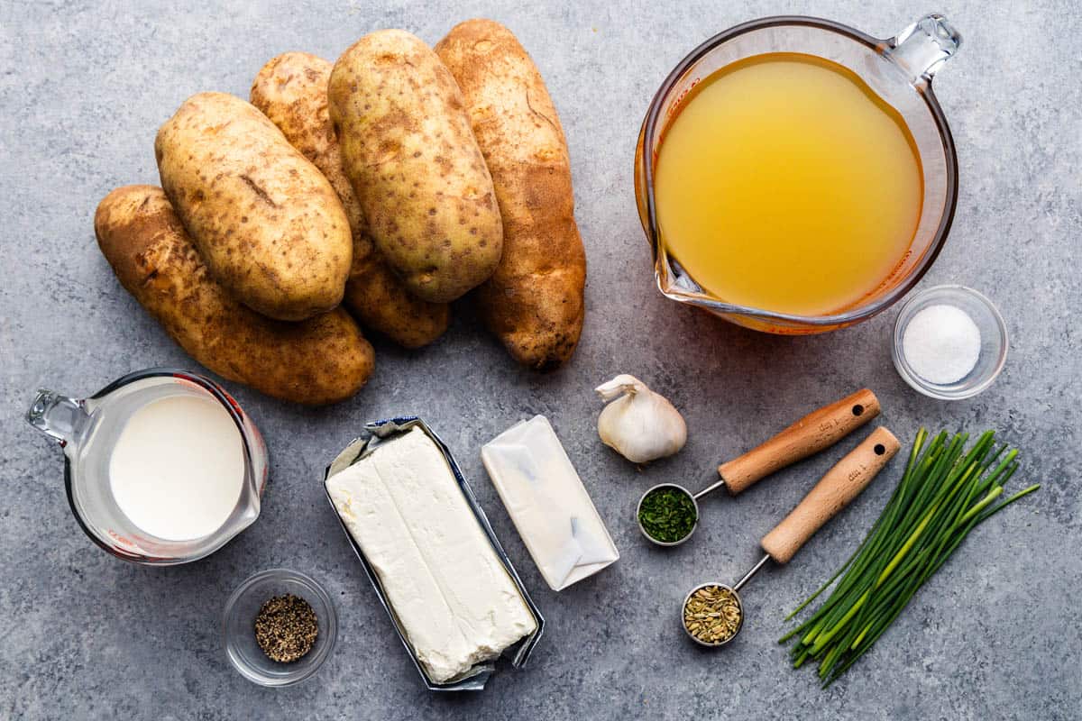 An overhead view of whole unpeeled potatoes next to measuring cups and spoons holding wet and dry ingredients needed to make herbed butter mashed potatoes.