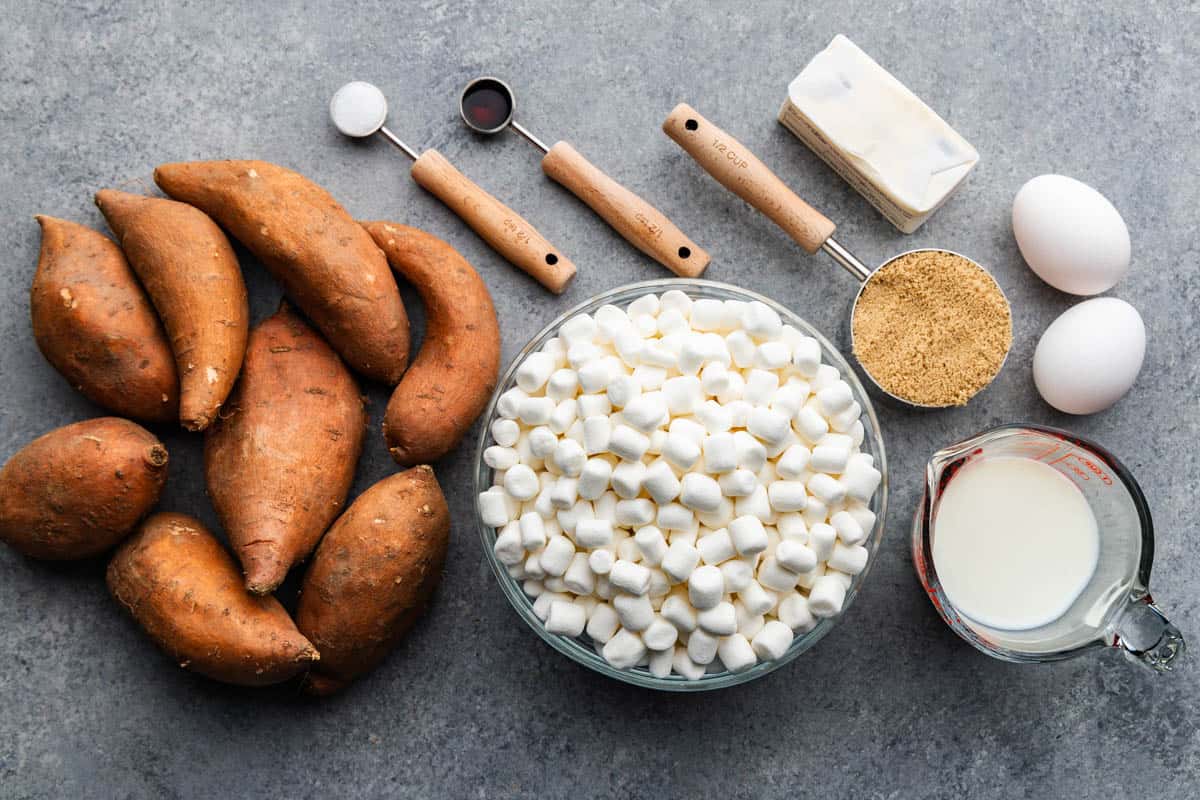 An overhead view of whole sweet potatoes next to bowls and measuring cups and spoons filled with ingredients to make sweet potato casserole with marshmallows.