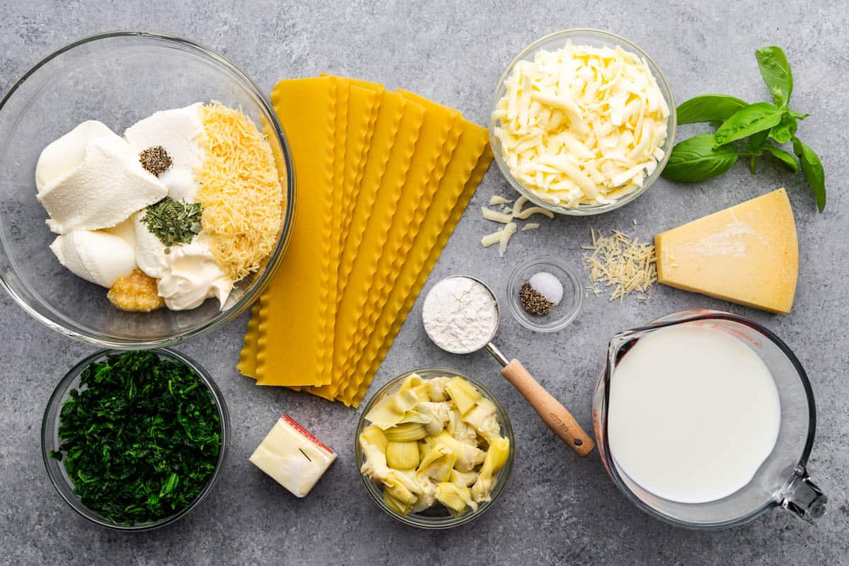 An overhead shot of fresh, wet, and dry ingredients needed to make white spinach artichoke lasagna.