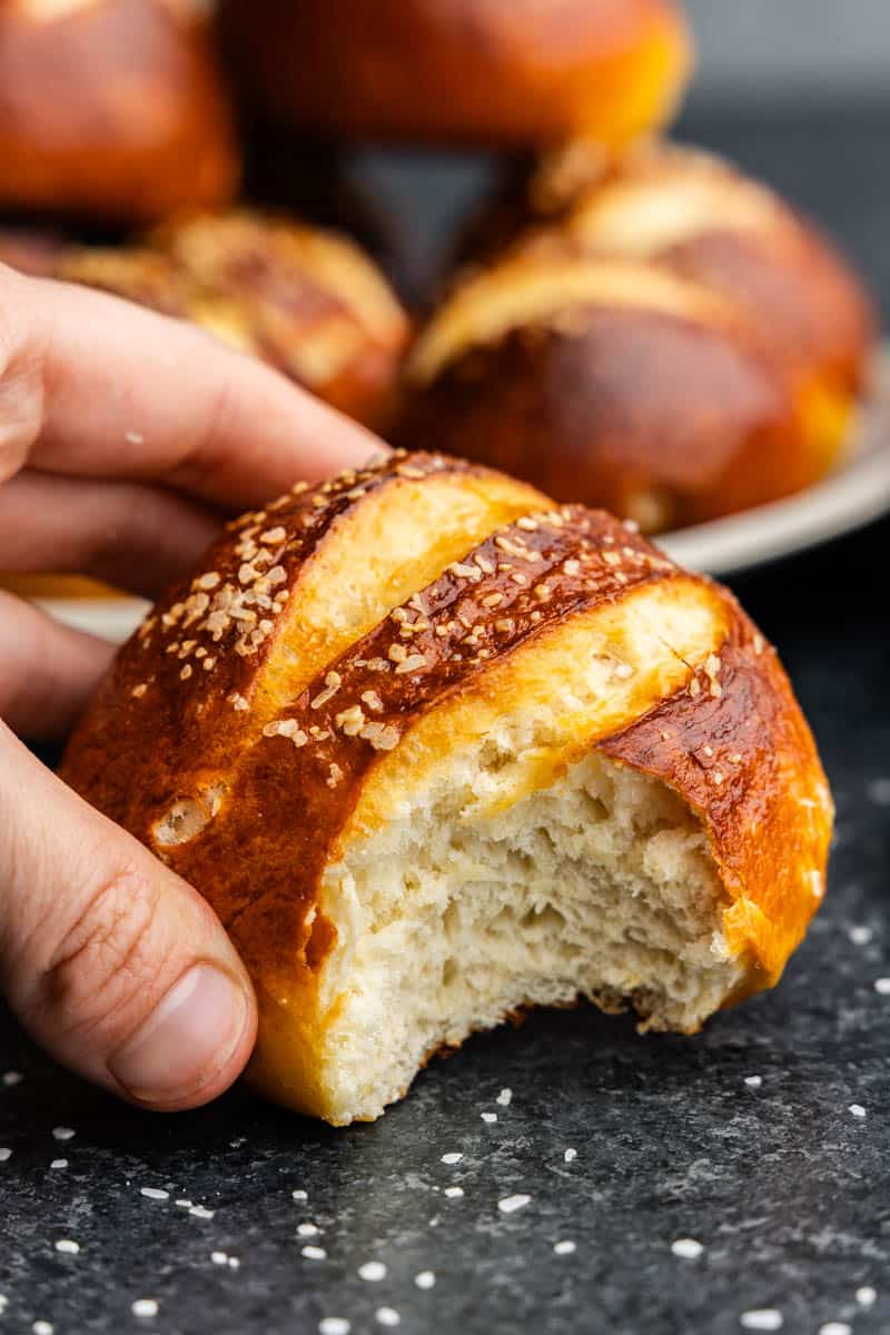 A close-up view of a hand holding a homemade pretzel roll from which a bite has been taken