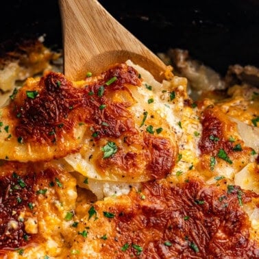 A close up view of scalloped potatoes made in the slow cooker. There is a wooden spoon ready to scoop out the first helping.