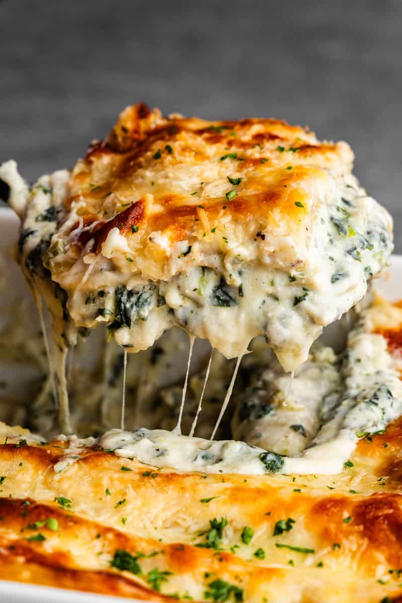 A square of white spinach artichoke lasagna being lifted from the pan with melted cheese stretching.