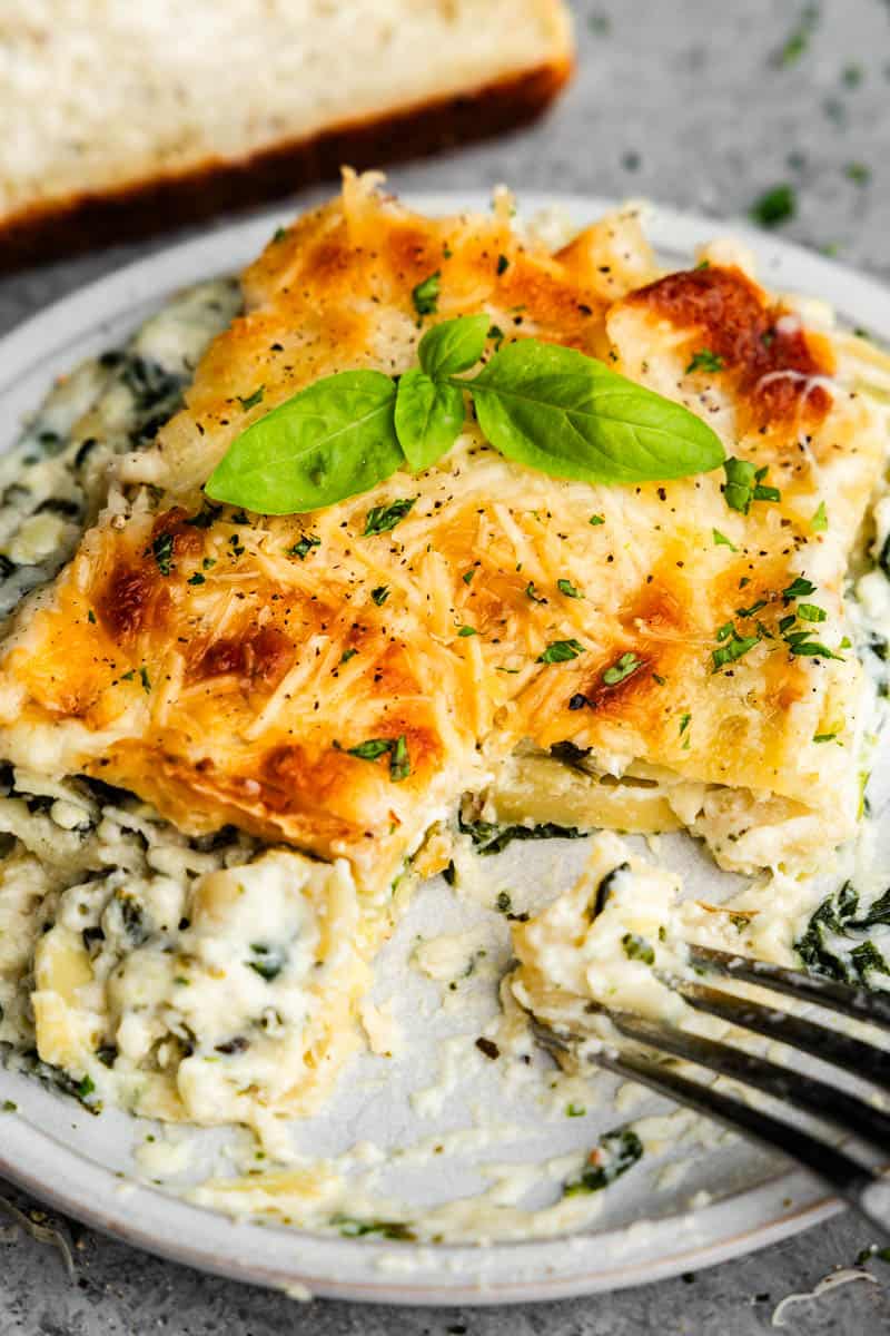 A close-up view of a square of white spinach artichoke lasagna on a plate with a bite taken out and a fork sitting in the corner of the plate.