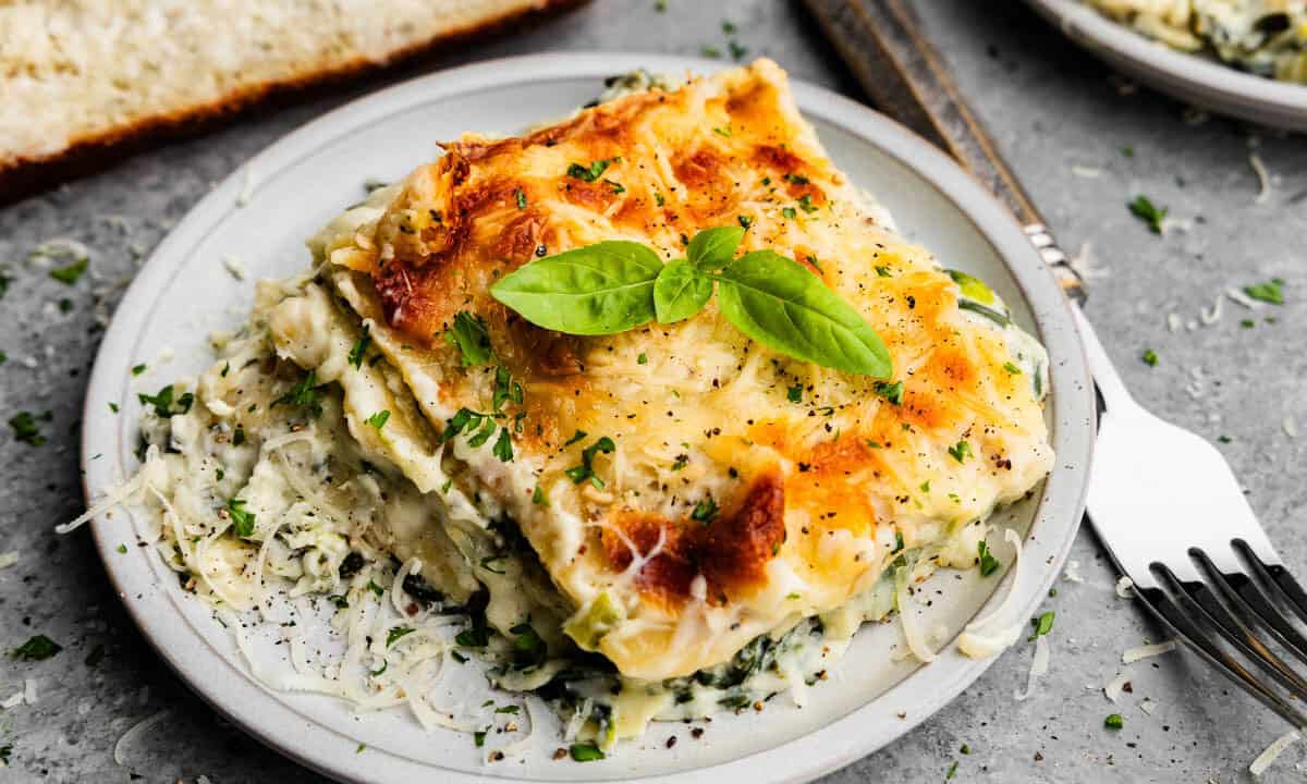 A square serving of white spinach artichoke lasagna on a plate set next to a fork, with fresh basil leaves on top.