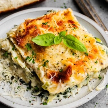 A square serving of white spinach artichoke lasagna on a plate set next to a fork, with fresh basil leaves on top.