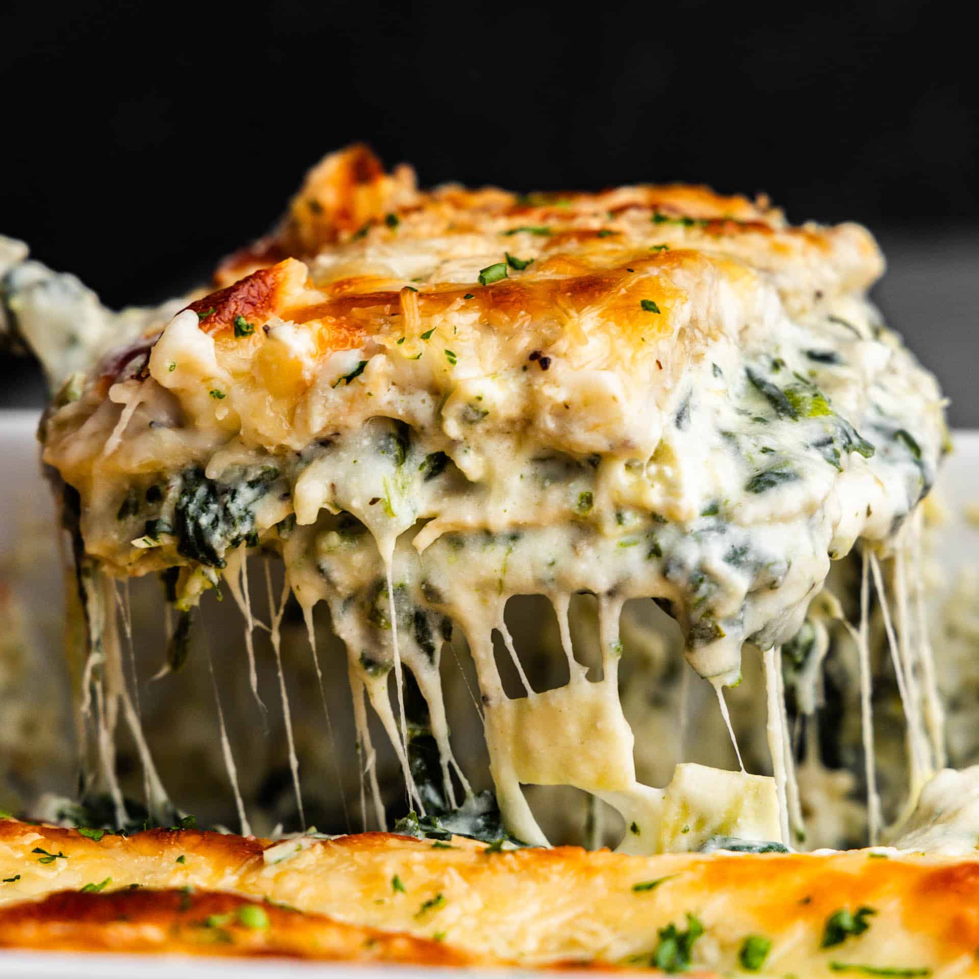 A side view of a spatula lifting a square of white spinach artichoke lasagna showing off the melty and stretchy cheese.