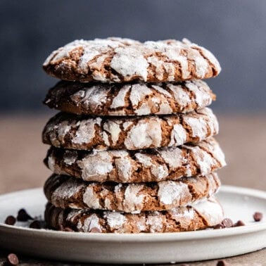 A plate with a tower of chocolate crinkle cookies place on it, surrounded by a scatter of mini chocolate chips.