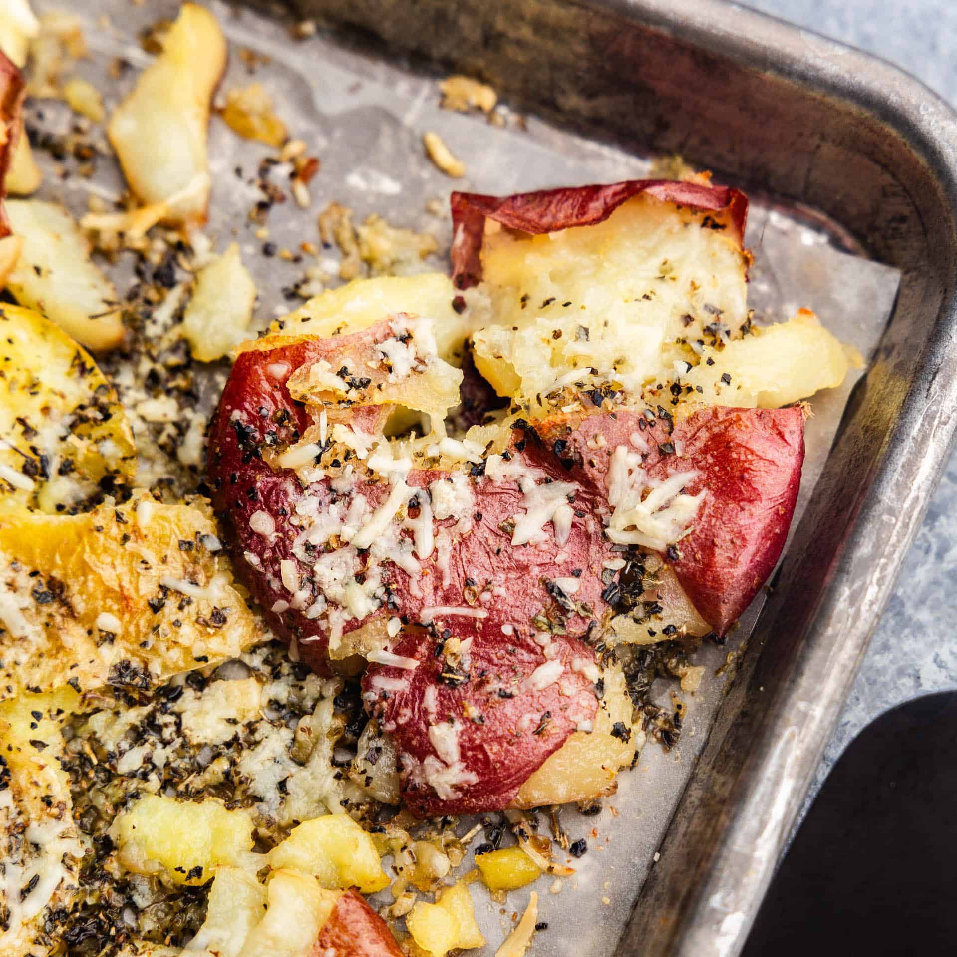 A close up of a smashed red potato on a baking sheet, baked golden brown and sprinkled with spices and parmesan cheese.