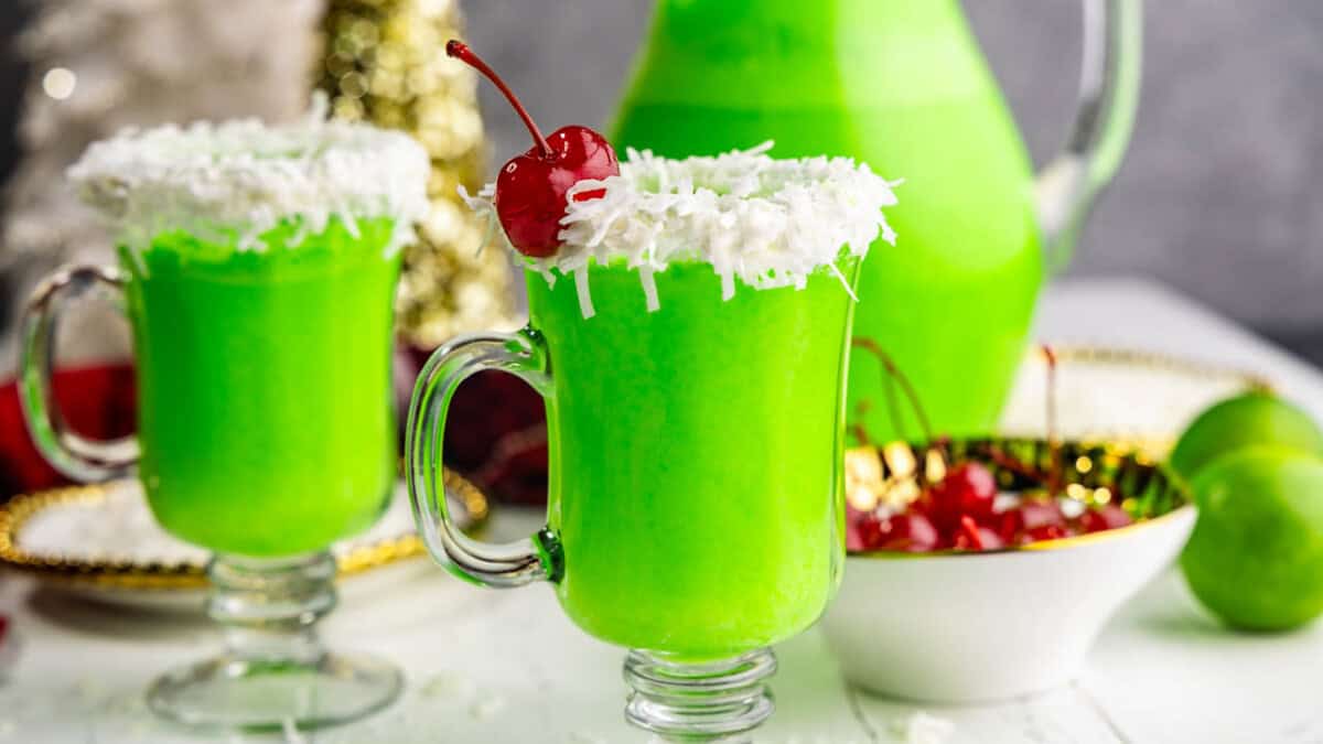 Two glasses of green grinch punch in a clear glass mug, with a white frosting and coconut rim and red cherry garnish.