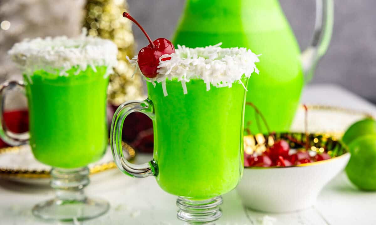 Two glasses of green grinch punch in a clear glass mug, with a white frosting and coconut rim and red cherry garnish.