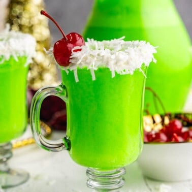 Two glasses of green grinch punch in a clear glass mug, with a white frosting and coconut rim and red cherry garnish.