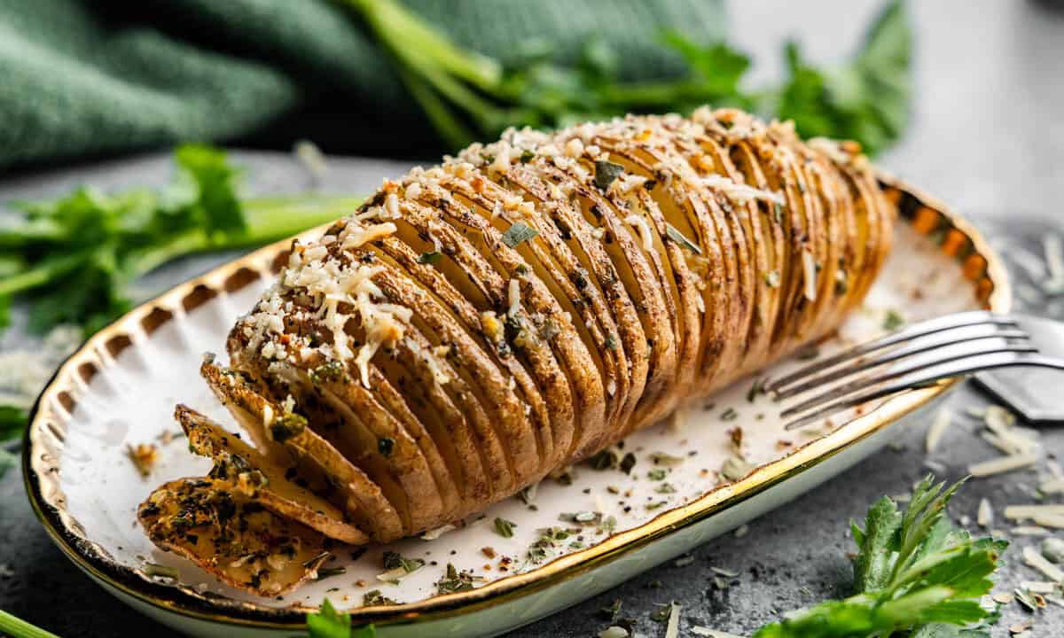 A hasselback potato on a plate with a fork resting on the edge of the plate.