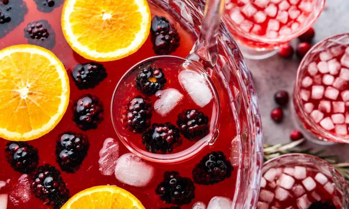 A close up view into a punch bowl filled with holiday punch, fresh blackberries and orange slices, with a ladle scooping out a serving.