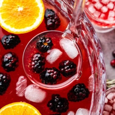 A close up view into a punch bowl filled with holiday punch, fresh blackberries and orange slices, with a ladle scooping out a serving.