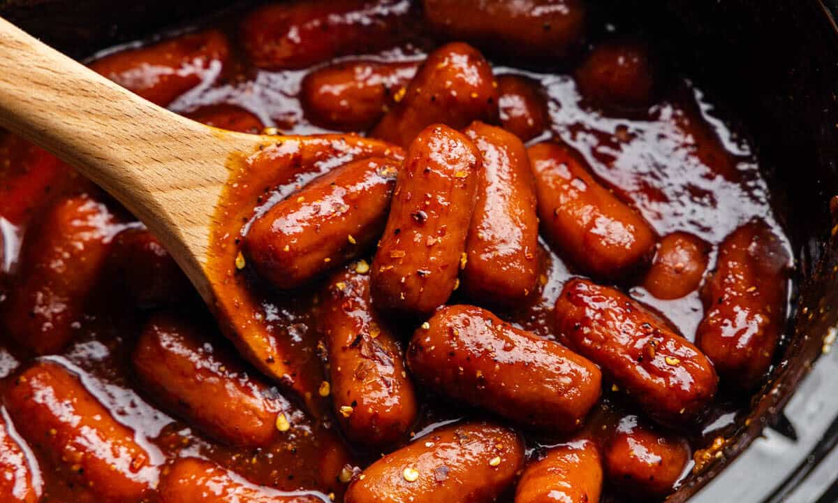 A close up view of lil smokies in a crockpot with a wooden spoon sticking out.