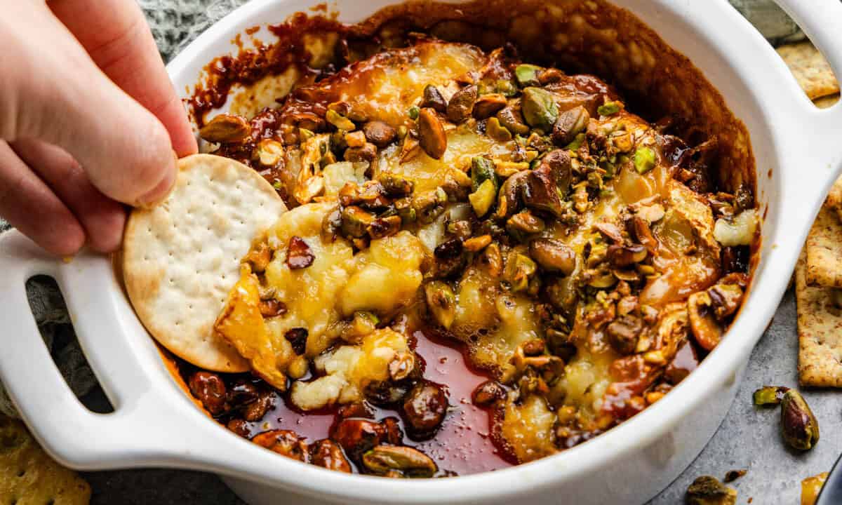 A hand dipping a chip into a baking dish with hot honey pistachio baked brie.