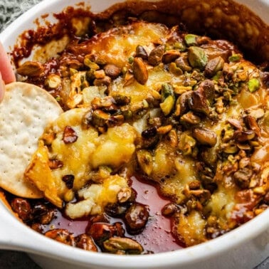 A hand dipping a chip into a baking dish with hot honey pistachio baked brie.