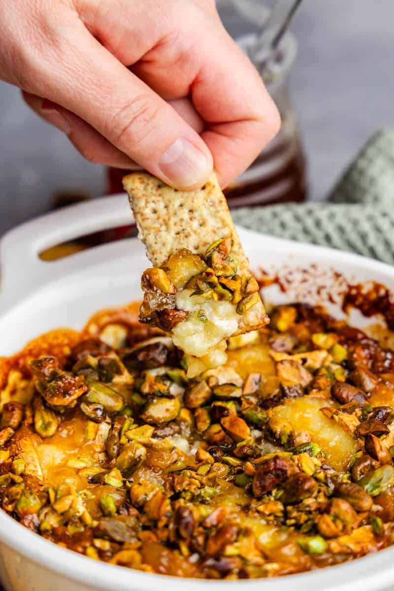 Fingers holding a cracker that is scooping out a bite of hot honey pistachio baked brie.