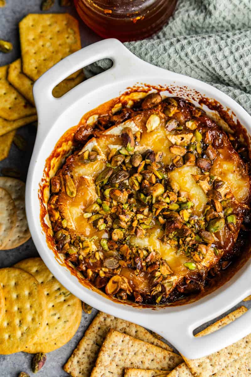 An overhead view of a white ceramic dish holding baked brie topped with hot honey and pistachios, with crackers set next to the bowl.