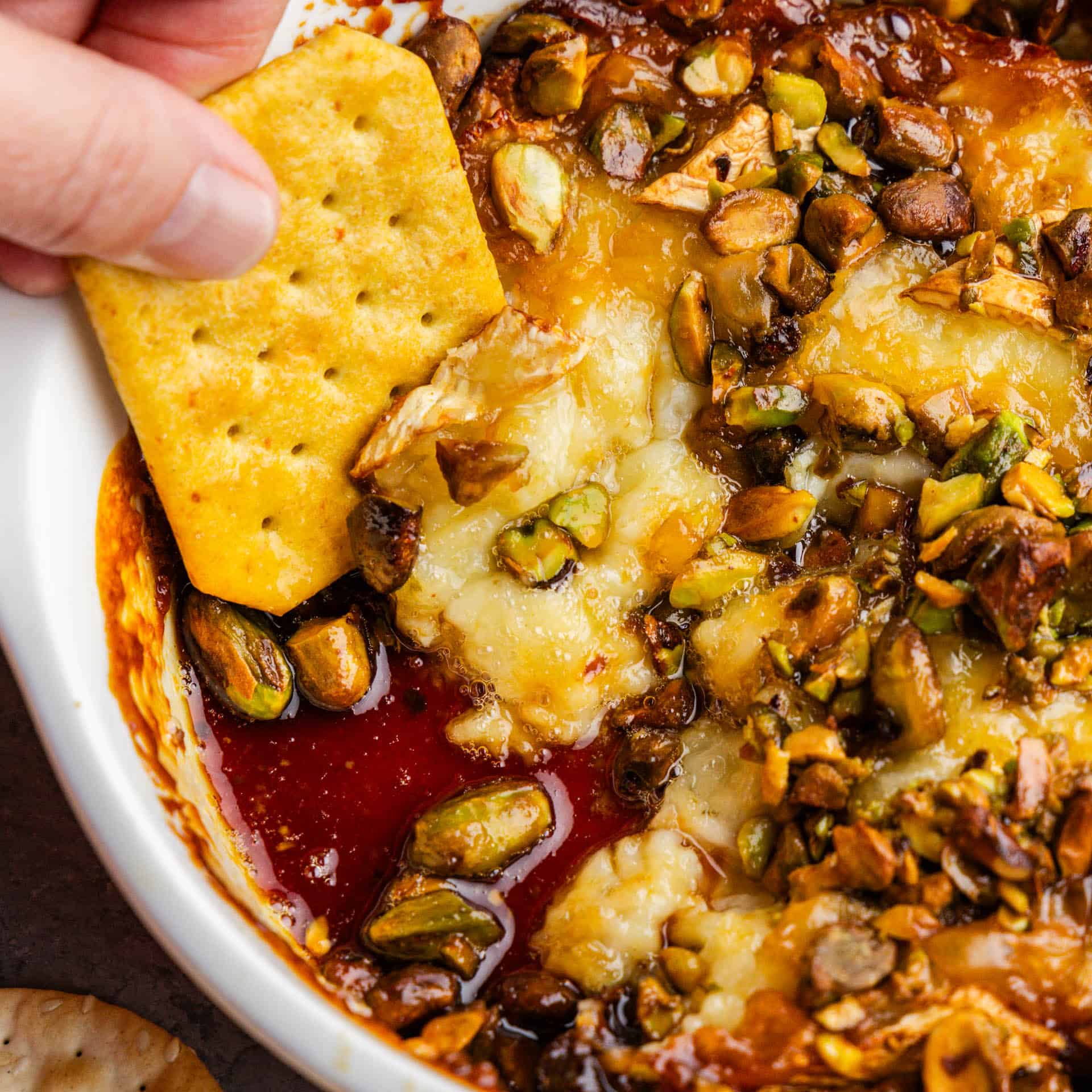 A close up of a cracker dipping into a bowl of hot honey pistachio baked brie.