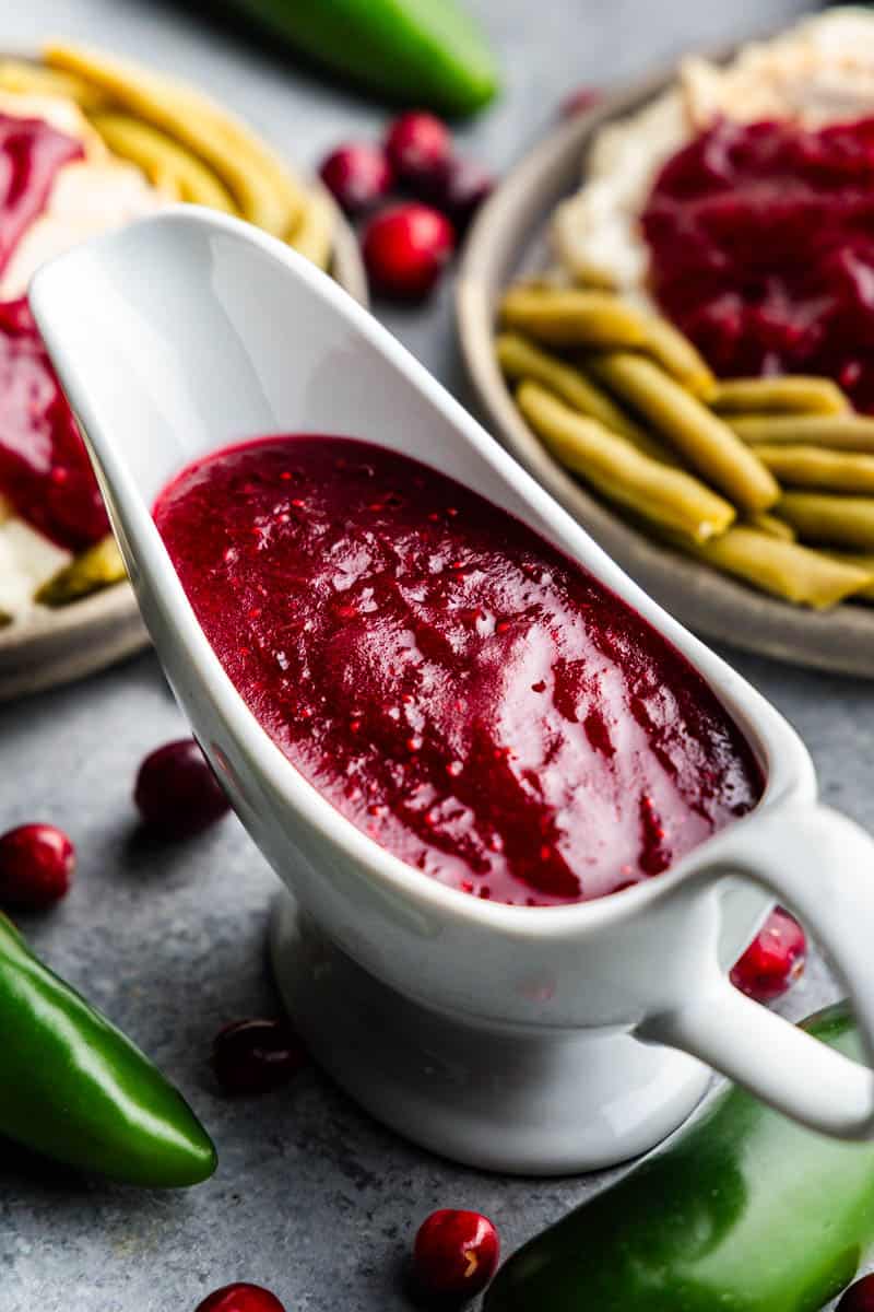 A close up overhead view of a white gravy boat filled with jalapeno cranberry sauce set on a counter next to a plates holding green beans, mashed potatoes and turkey, covered in jalapeno cranberry sauce.