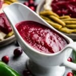 A white ceramic gravy boat filled with jalapeno cranberry sauce, set on a counter amongst scattered fresh cranberries and a whole jalapeno, with plates in the background holding servings of turkey topped with jalapeno cranberry sauce and green beans.