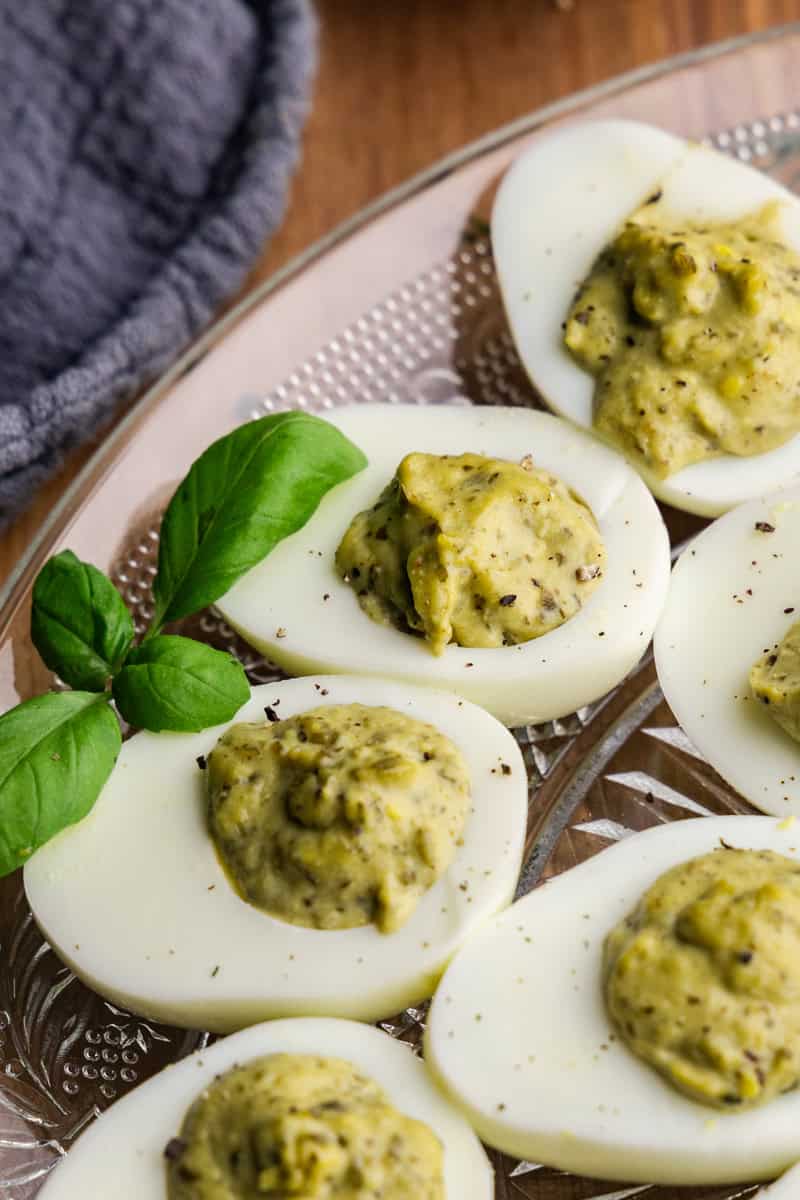 A close up of pesto deviled eggs on a glass serving plate with a fresh sprig of basil.