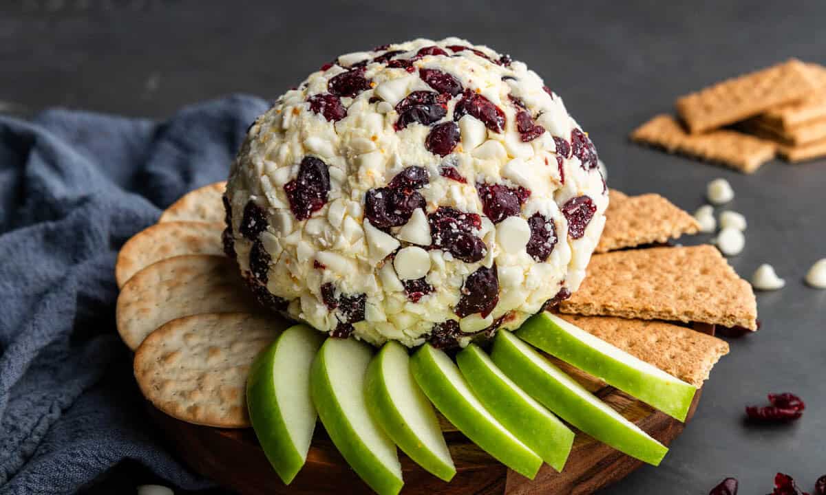 A white chocolate cranberry orange cheese ball surrounded with sliced green apples, crackers and graham crackers.