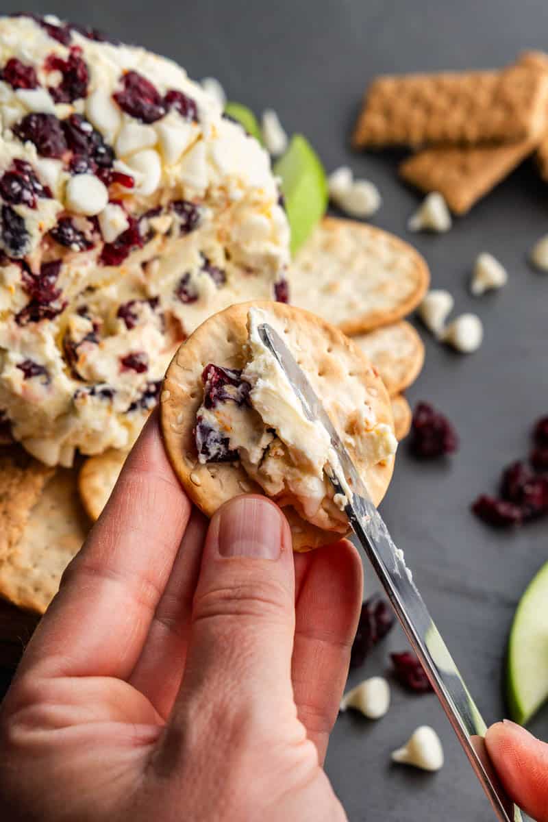 A hand holding a cracker while spreading white chocolate cranberry orange cheese ball onto it with a knife.