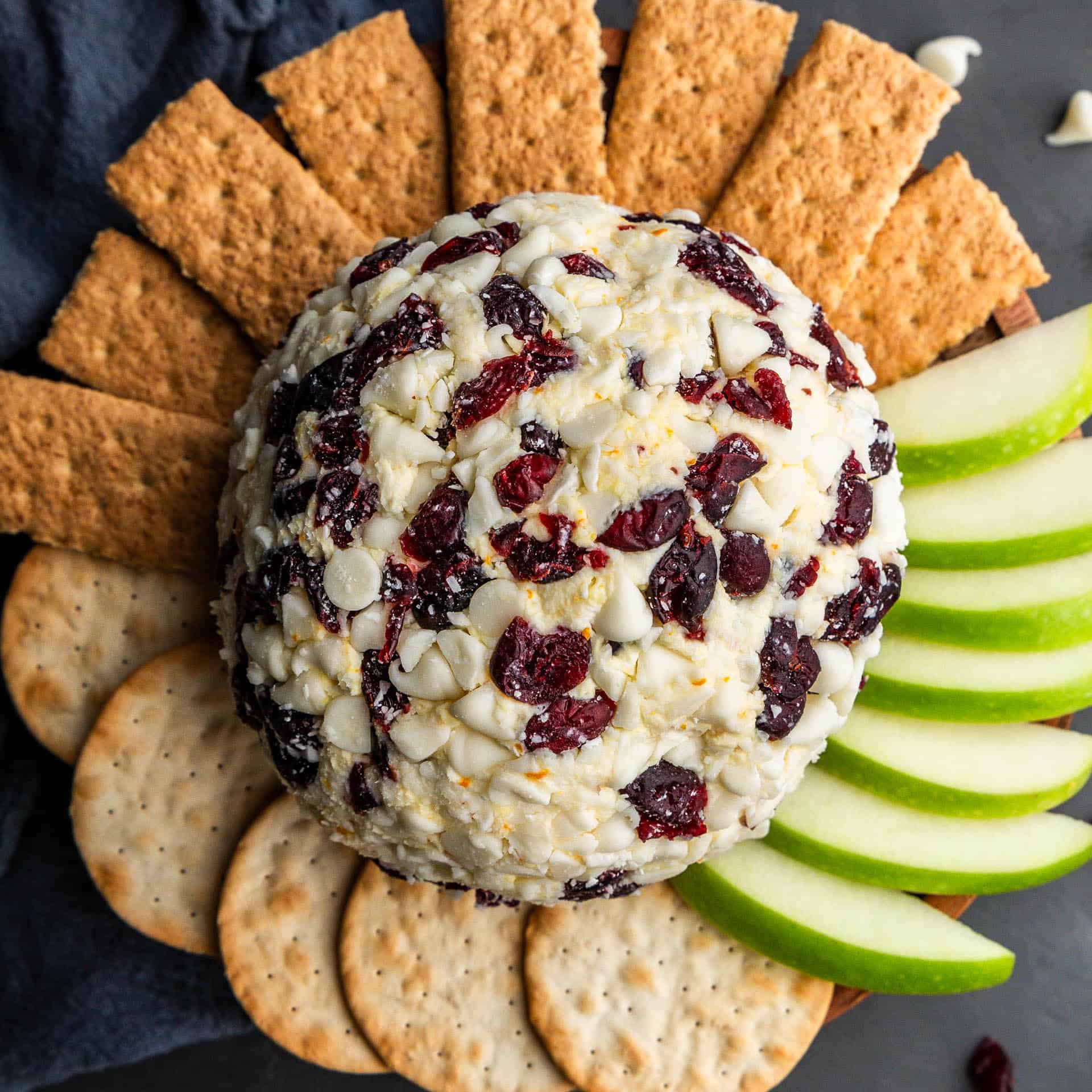 An overhead view of a white chocolate cranberry orange cheese ball surrounded by crackers, apples, and graham crackers that are fanned out.