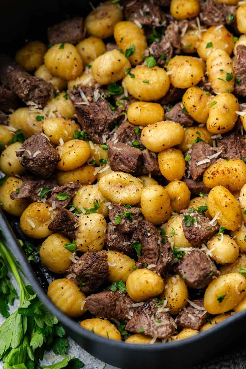 A close up overhead view of steak bites and gnocchi in the basket of an air fryer.