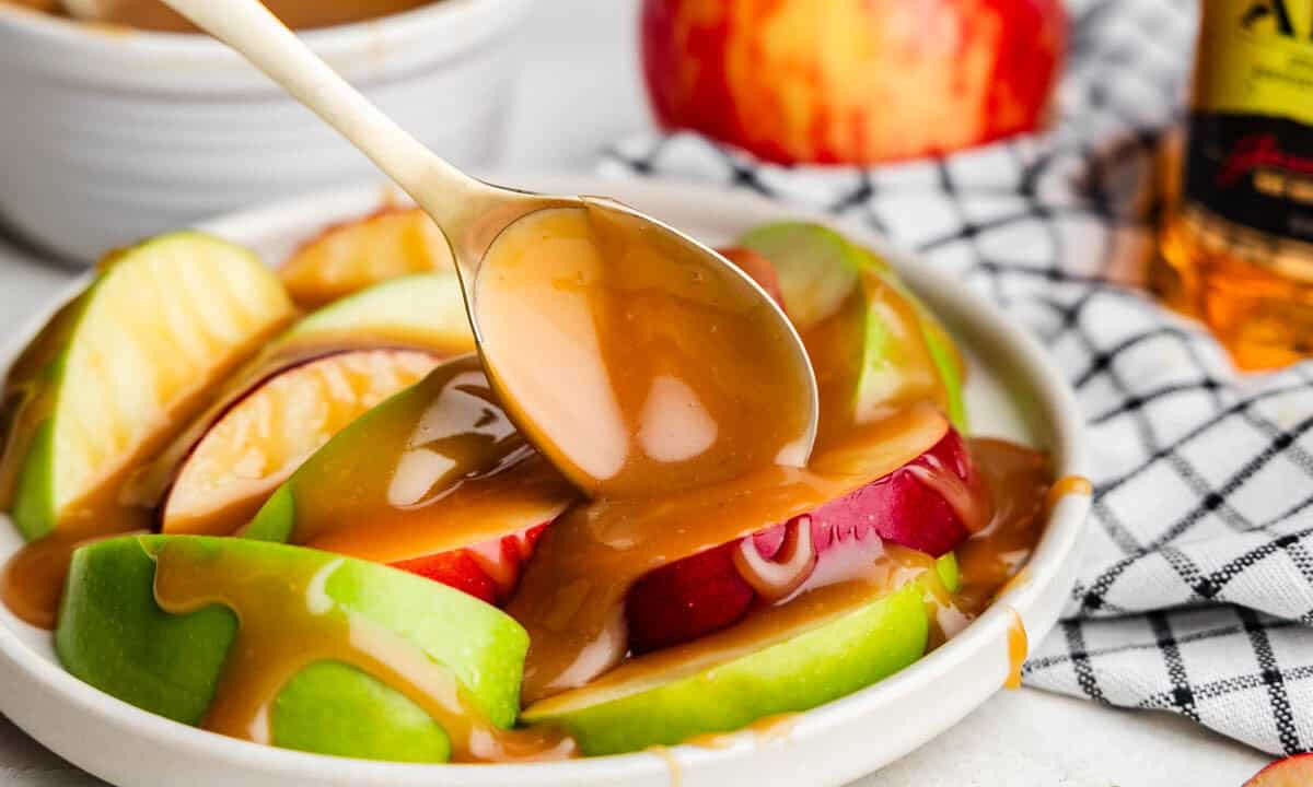 A spoon drizzling apple bourbon caramel sauce over a plate of sliced apples.