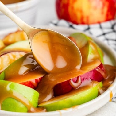 A spoon drizzling apple bourbon caramel sauce over a plate of sliced apples.