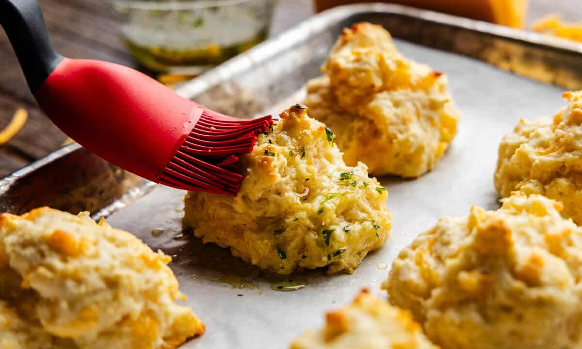 A silicone brush brushing butter onto a cheddar bay biscuit on a pan.