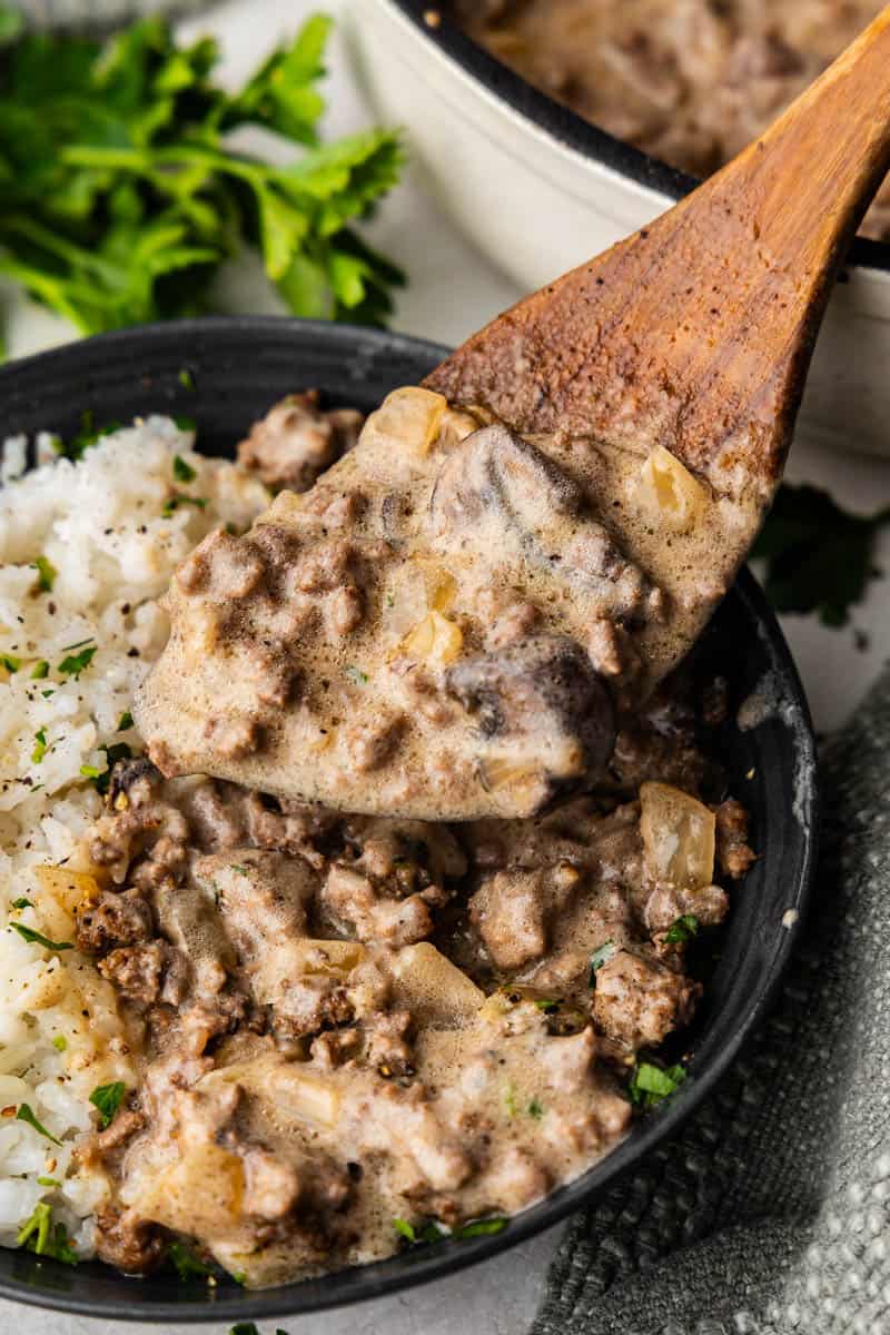 A wooden spoon scooping a helping of ground beef stroganoff and pouring it over white rice.