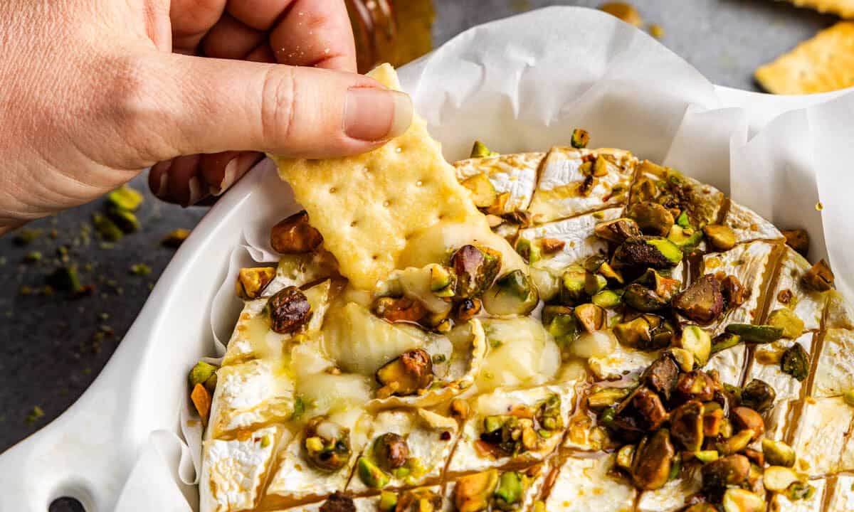 A cracker being dipped into hot honey pistachio baked brie.