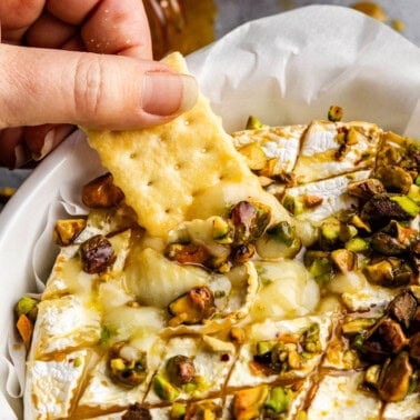 A cracker being dipped into hot honey pistachio baked brie.