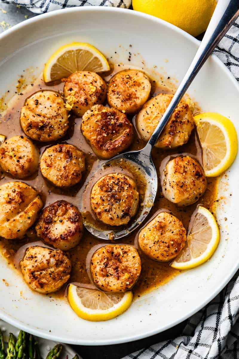 An overhead view of a white plate with lemon garlic scallops and lemon wedges.