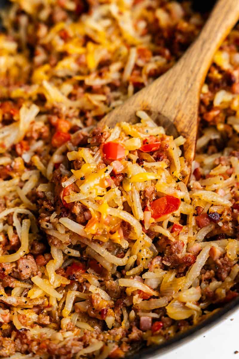 A close up of a wood spoon scooping out some loaded breakfast hash from the skillet.