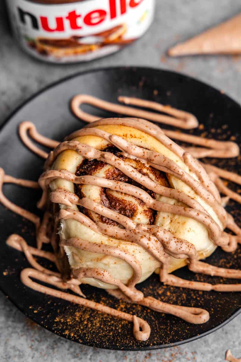 A Nutella cinnamon roll on a black plate with Nutella frosting generously drizzled all across the top.